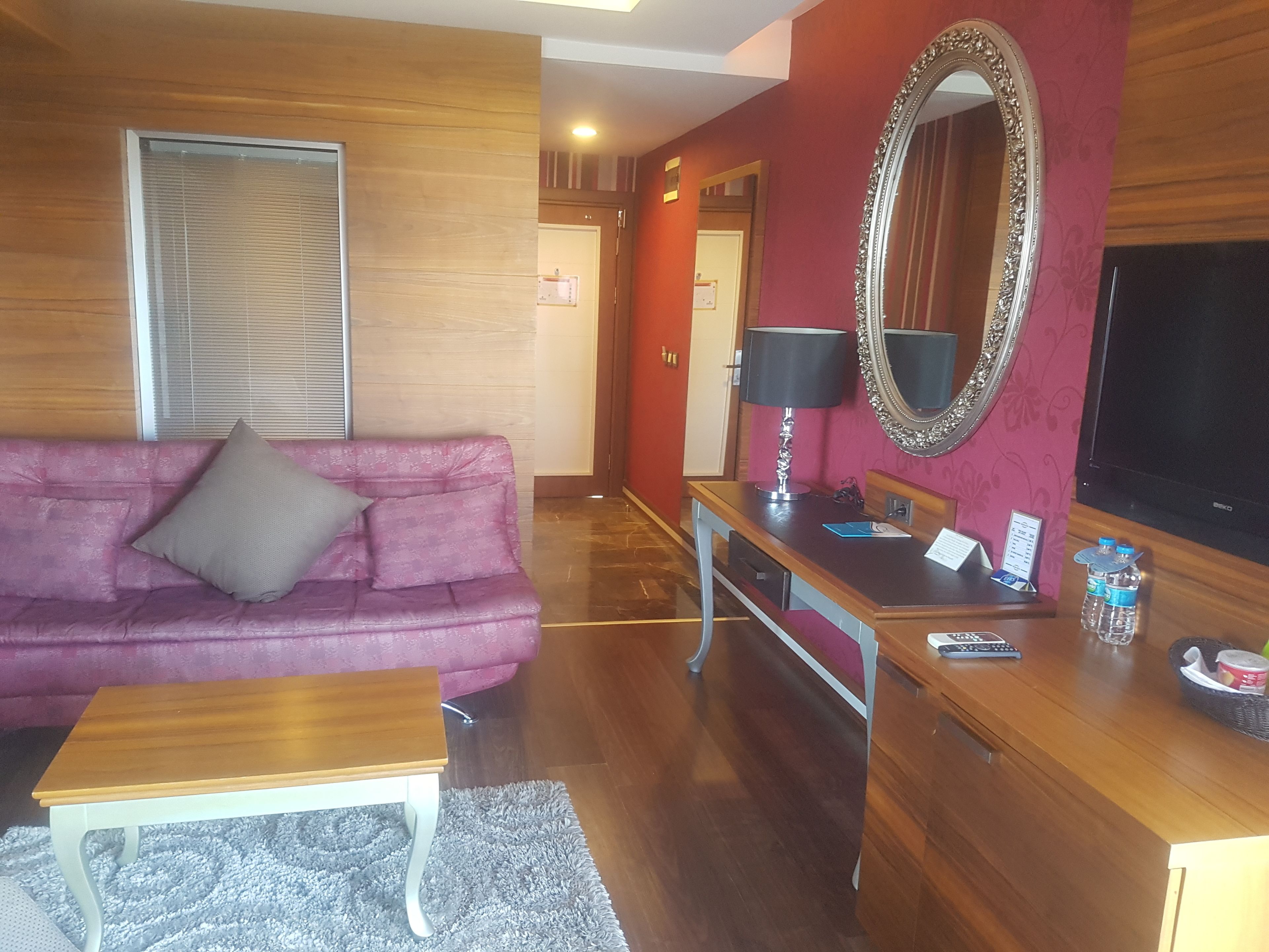 A living room with a wooden coffee table, a pink sofa, a wooden desk with a lamp, a large mirror, and a flat-screen TV.