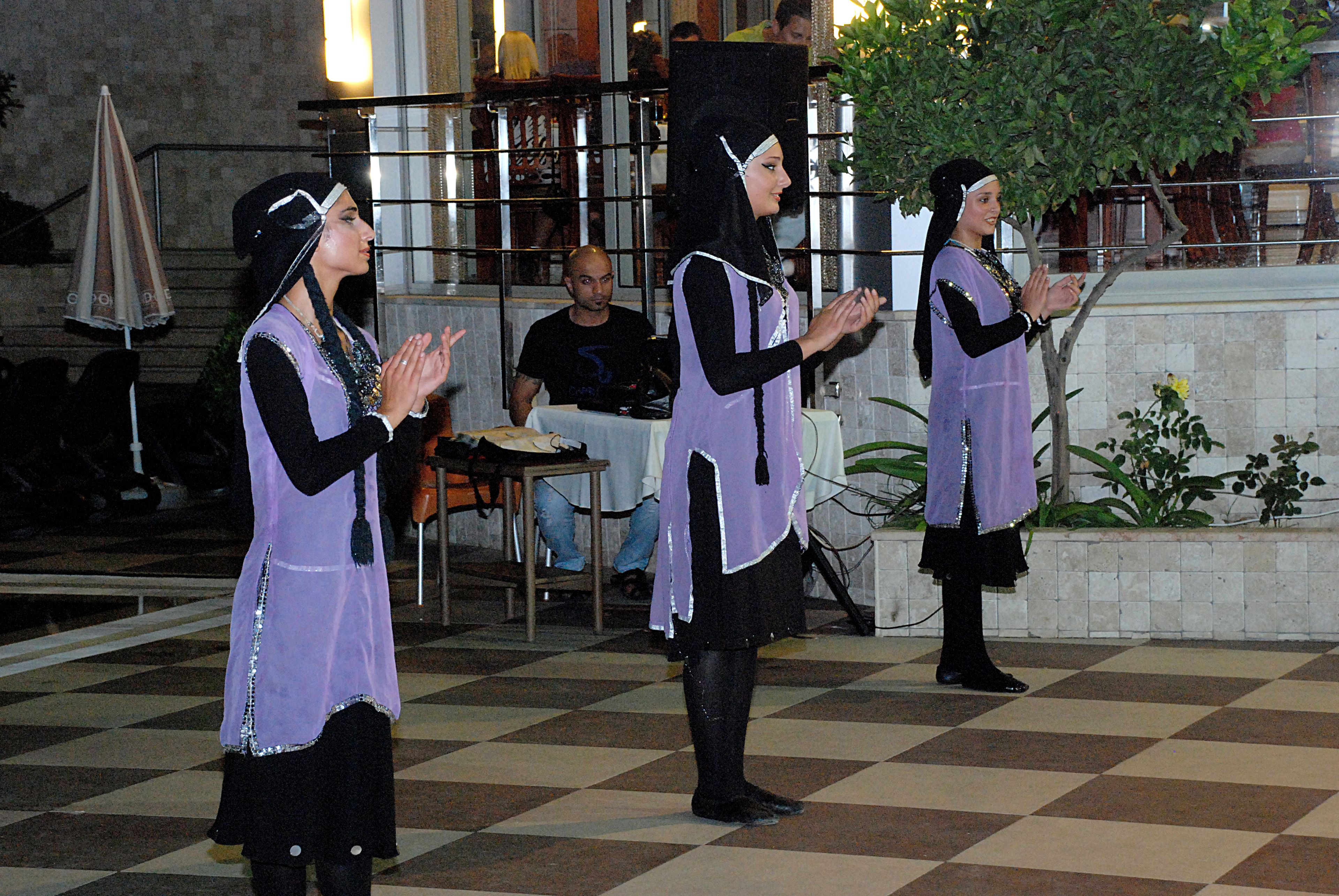 A group of people in traditional attire performing in an outdoor setting with a checkered floor and a building in the background.