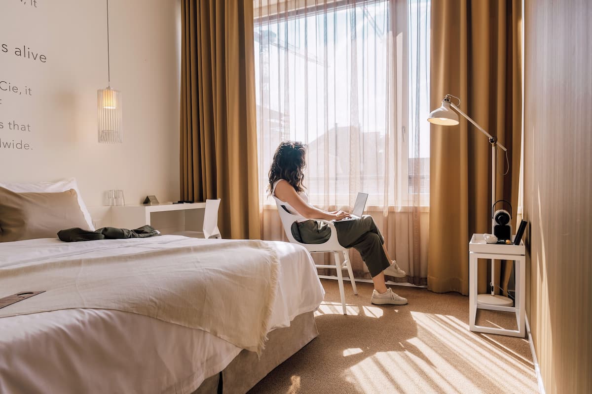 A hotel room with a bed, a desk, a chair, and a laptop.