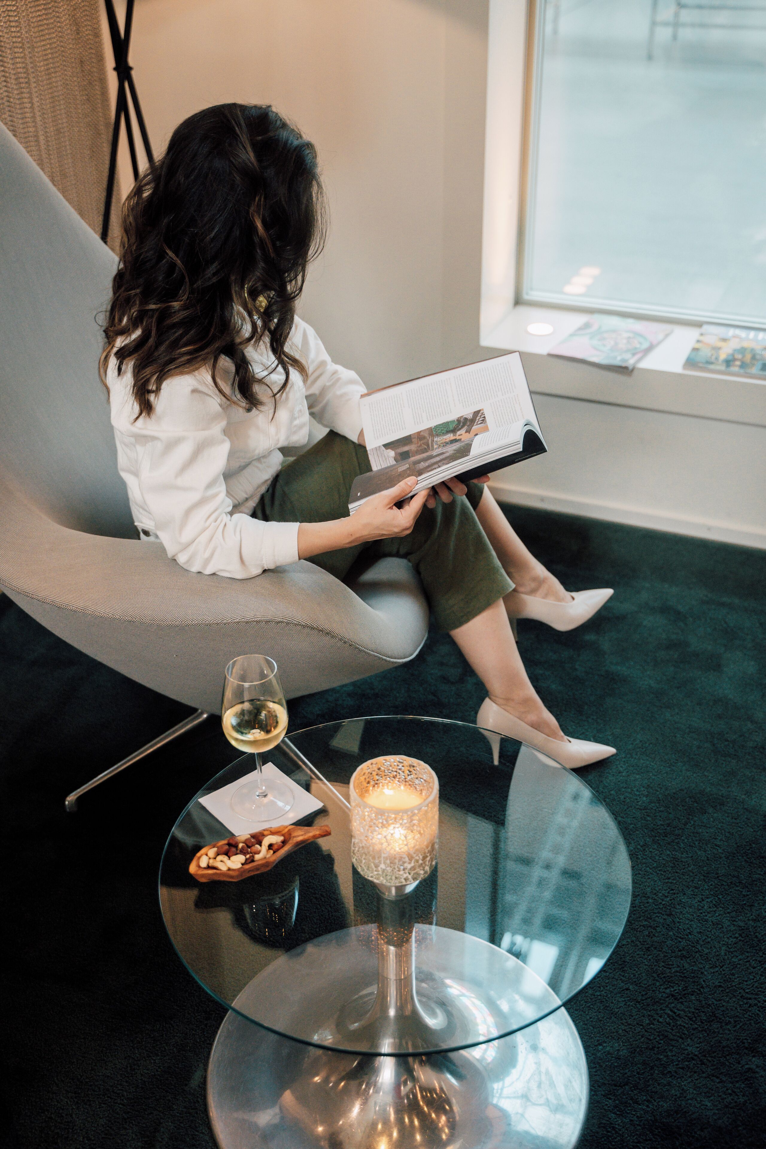 A woman sitting on a grey armchair reading a magazine, with a glass of wine and a candle on a glass table beside her.