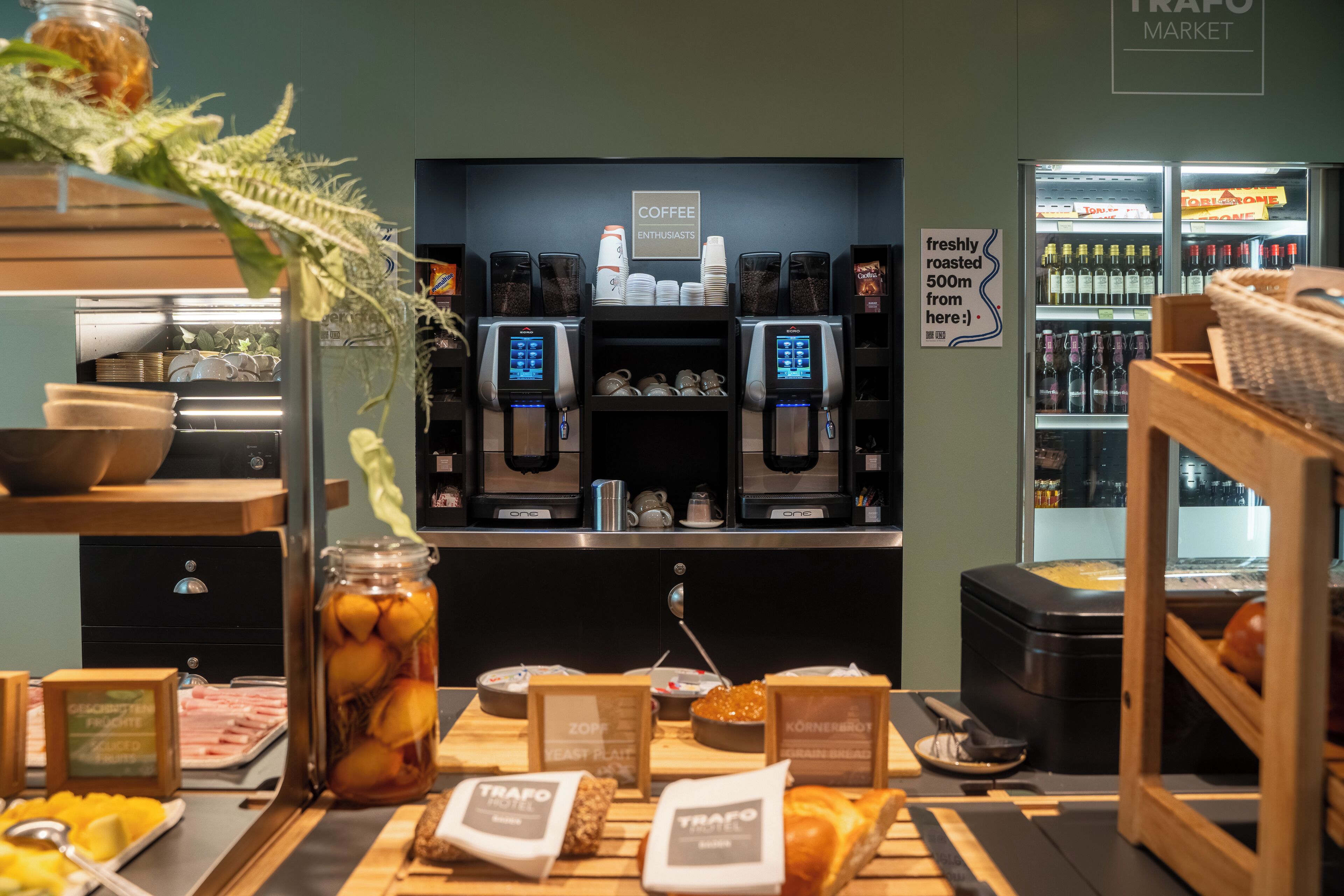 A coffee station with two espresso machines, a refrigerated display case with beverages, and a counter with food items.