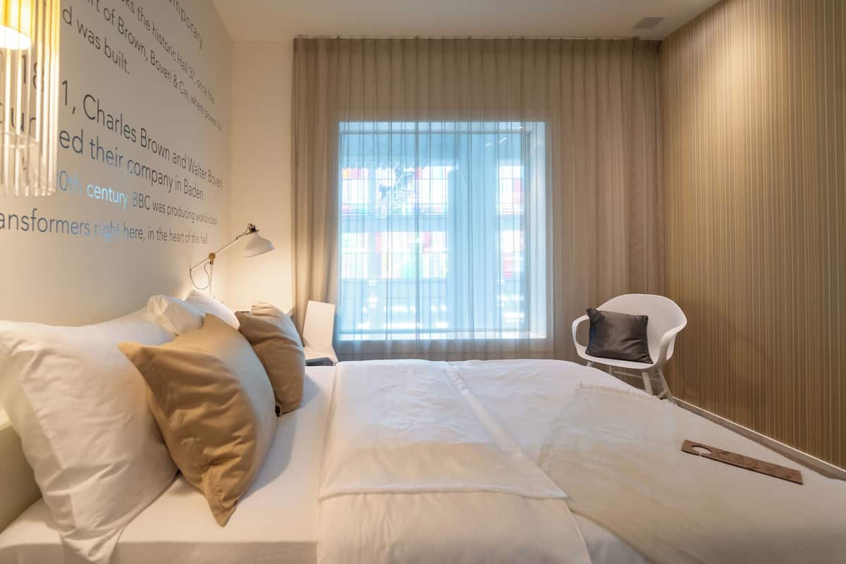 A hotel room with a large bed, a chair, a lamp, and a window with curtains.
