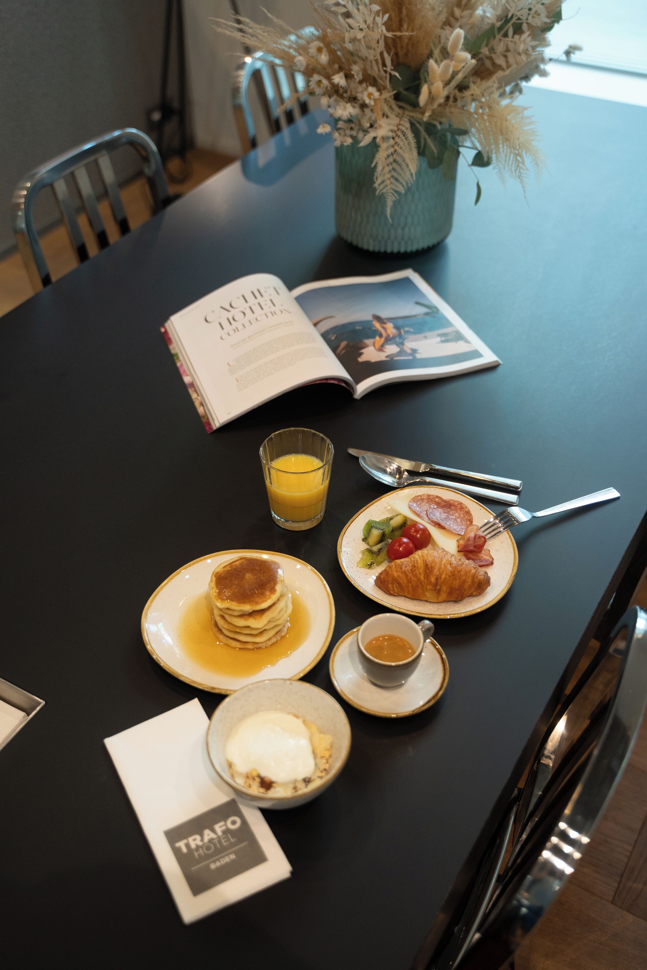 Une table dressée pour le petit-déjeuner, avec des crêpes, des croissants et une assiette de fruits, accompagnée d’un verre de jus d’orange, d’une tasse de café et d’un menu.