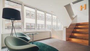 A modern interior with a staircase, a green chair, a lamp, and large windows. - Trafo Hotel (Baden)