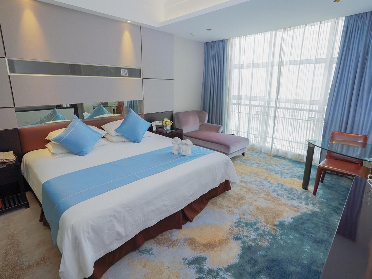Quarto de hotel com uma cama grande, uma escrivaninha, uma cadeira e vista da cidade por grandes janelas.