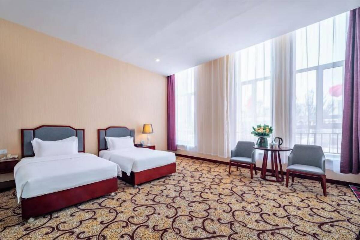 Hotellihuone, jossa on kaksi sänkyä, pieni pöytä tuoleineen, lamppu ja näkymä suurista ikkunoista.