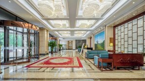Un vestíbulo de hotel con una zona de estar central, alfombra roja y un amplio ventanal con vistas a zonas verdes.