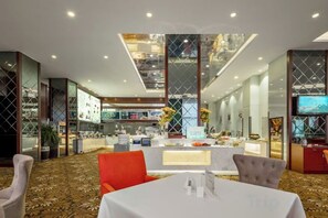 Un moderno vestíbulo de hotel con un mostrador de recepción central, un área de comedor con una mesa blanca y un gran ventanal con vista al interior.