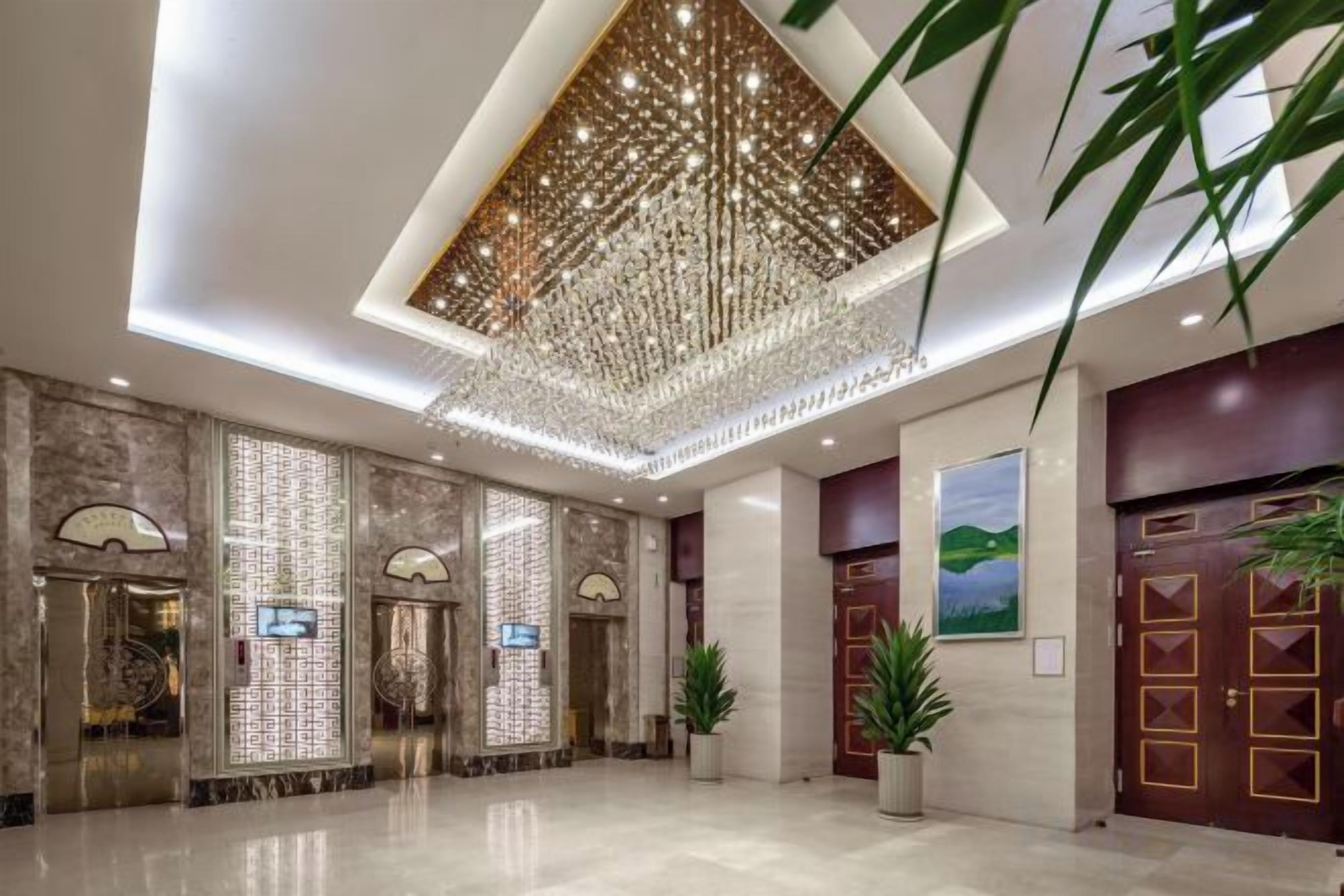 A hotel lobby with a chandelier, elevators, potted plants, and a painting on the wall.