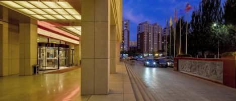 Entrada de hotel com design moderno, fachada de vidro e calçada pavimentada que leva à entrada.