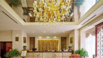 A hotel lobby with a reception desk, a chandelier, and potted plants.