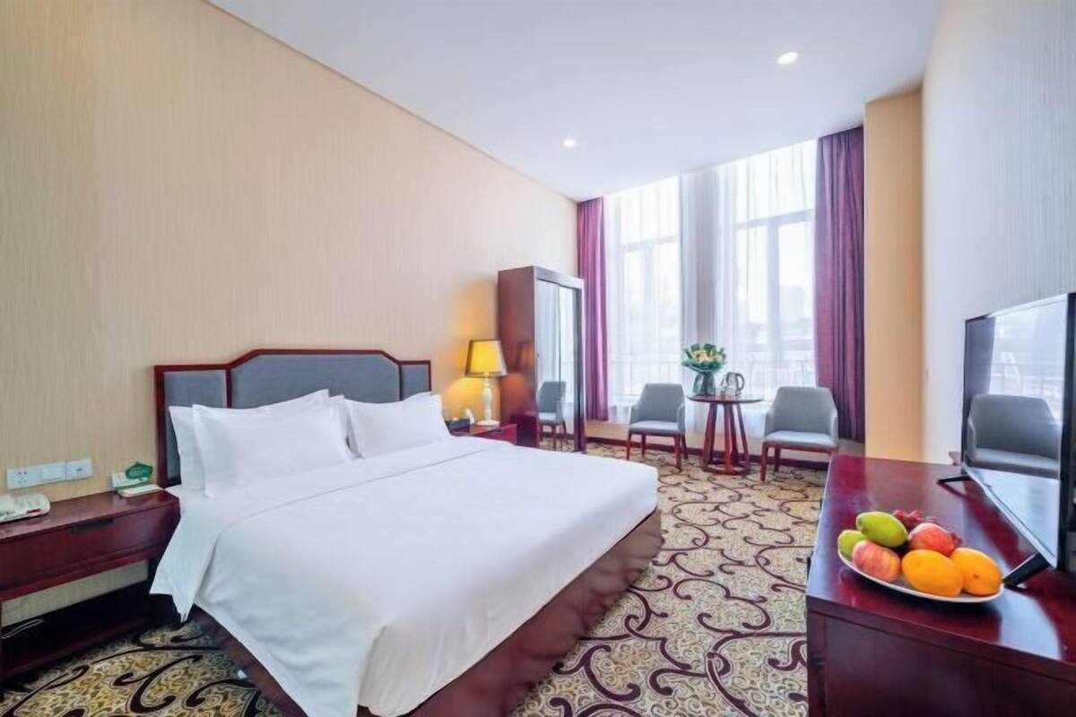 Hotellihuone, jossa on suuri sänky, työpöytä, jossa on televisio, pieni pöytä tuoleineen sekä hedelmäkulho.