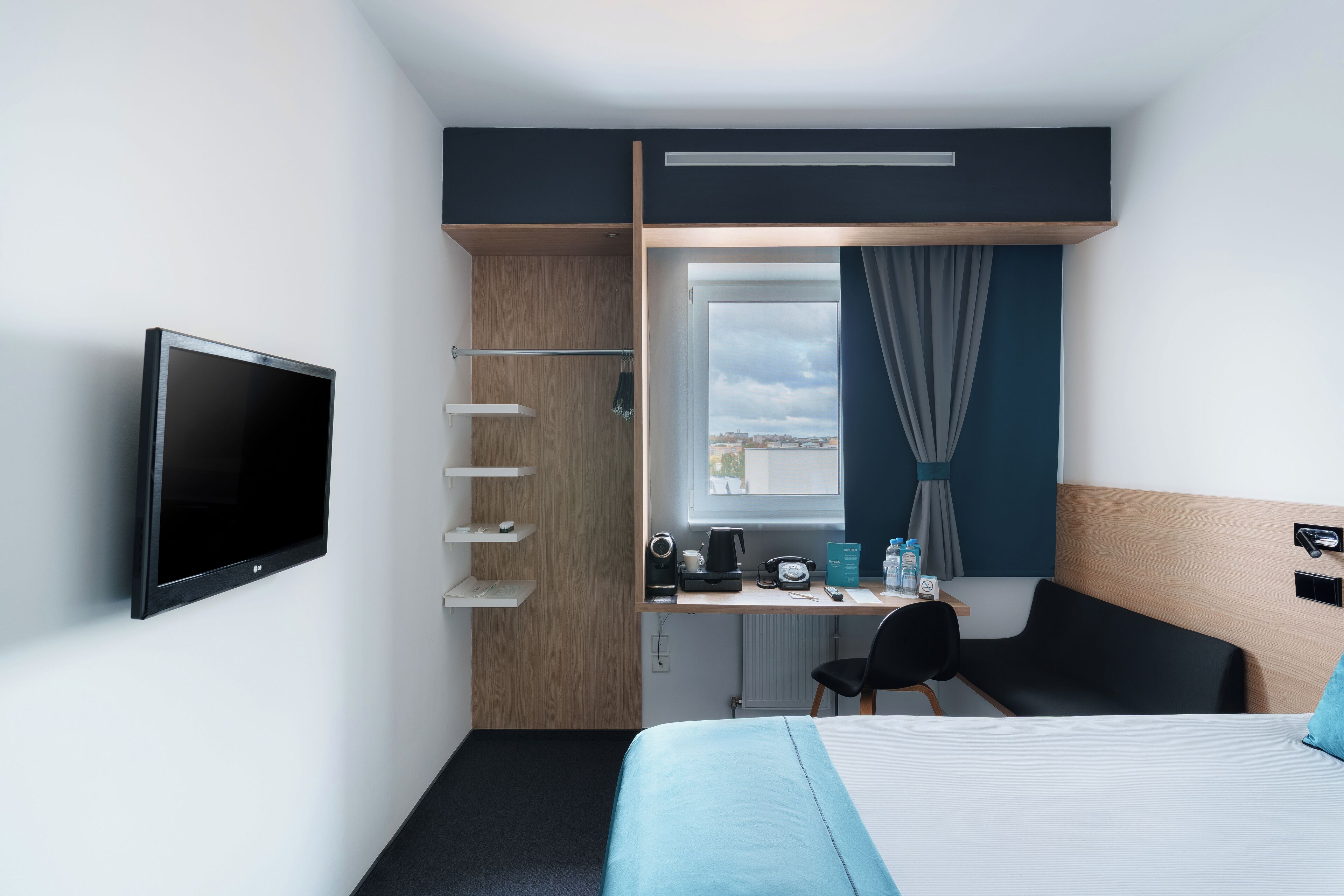 A modern hotel room with a bed, a desk with a computer, a TV, and a window with curtains.