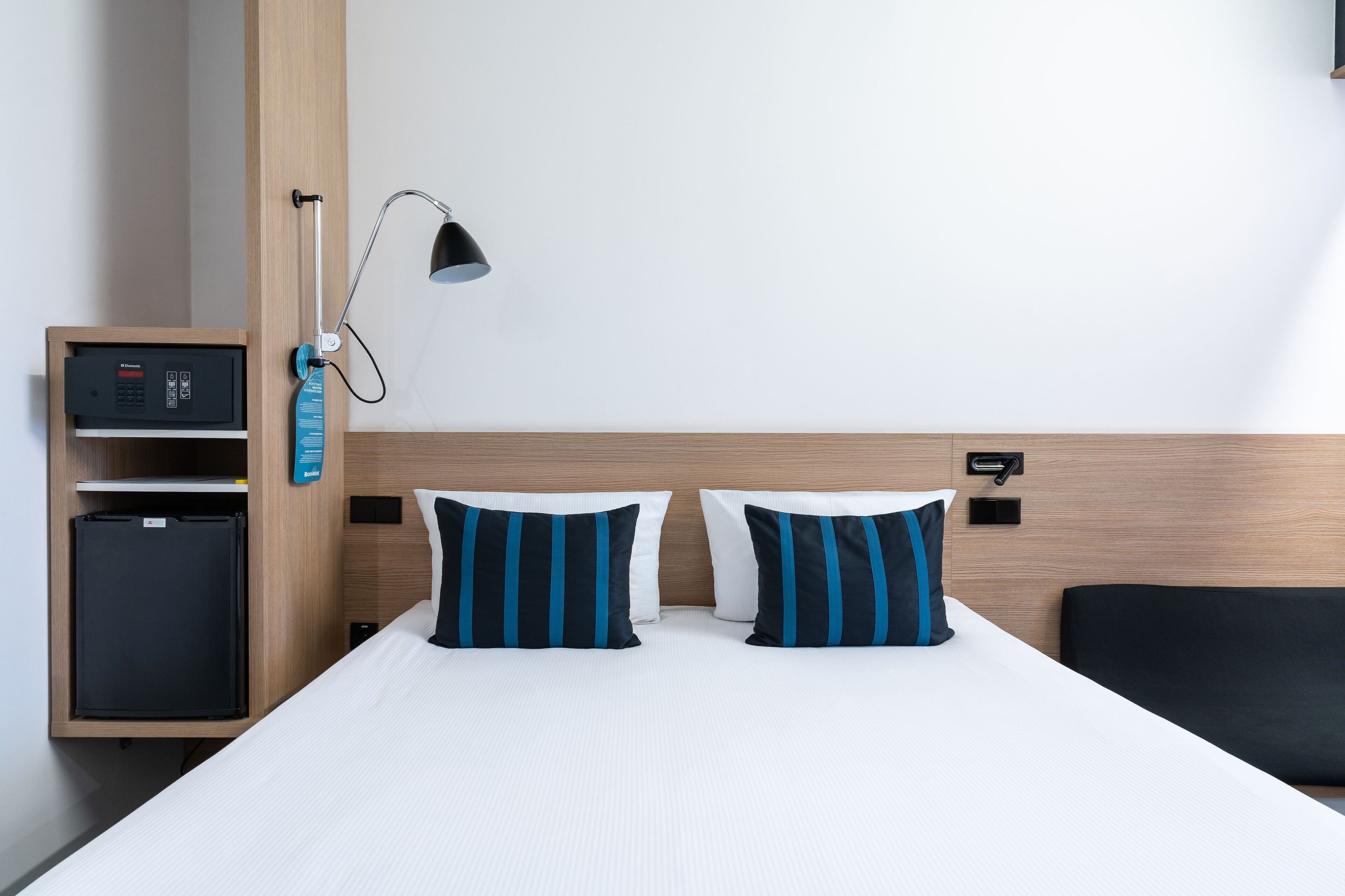 A hotel room with a bed, two pillows, a nightstand, a lamp, and a minibar.