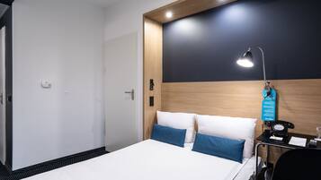 A hotel room with a bed, bedside table, lamp, and a wall-mounted telephone.
