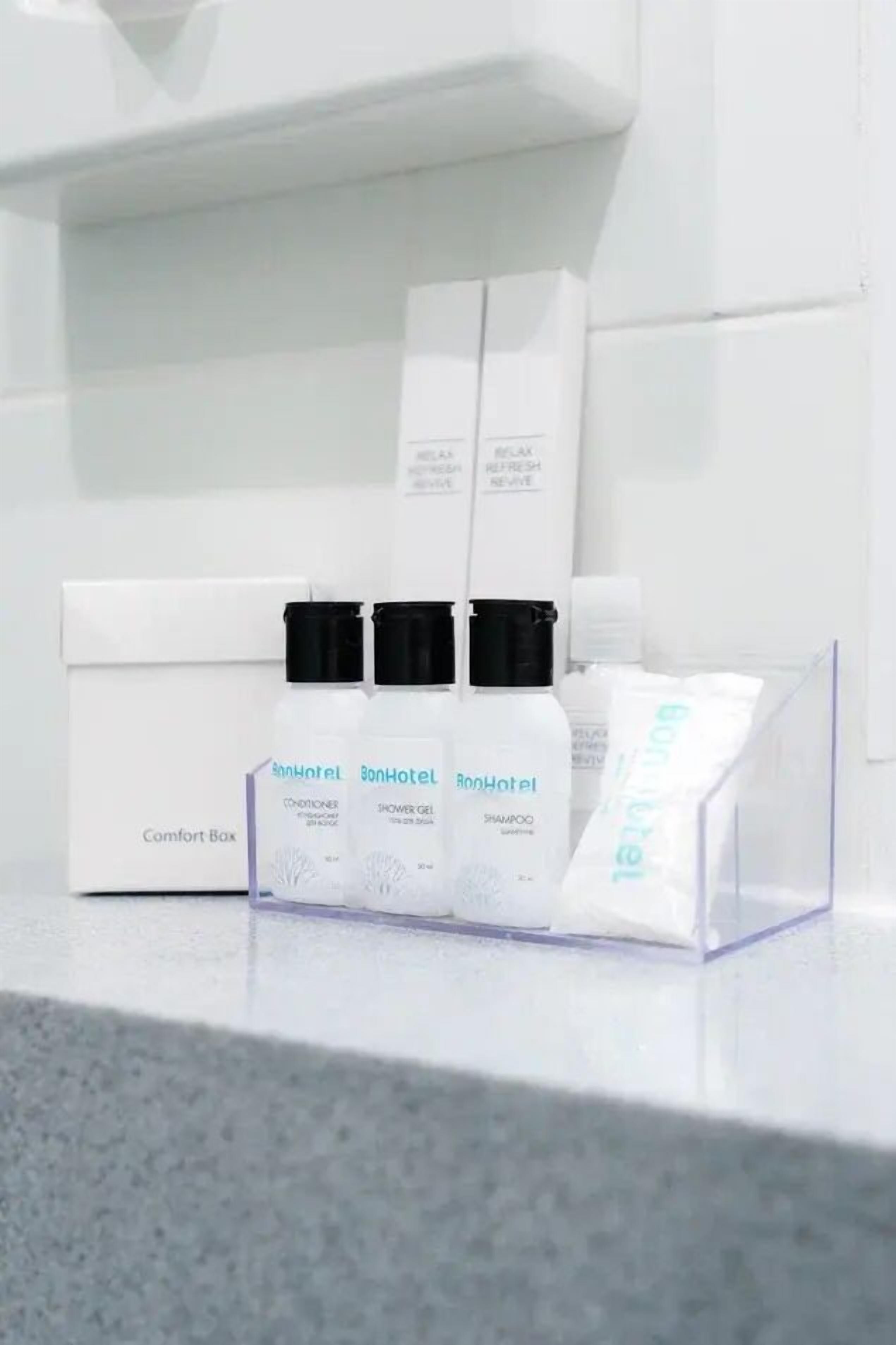 A hotel bathroom with a white sink, a mirror, and a shelf with toiletries including a conditioner, shower gel, and shampoo.