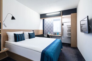 Una habitación de hotel con una cama grande, un escritorio con ordenador, un televisor montado en la pared y una ventana con un diseño geométrico.