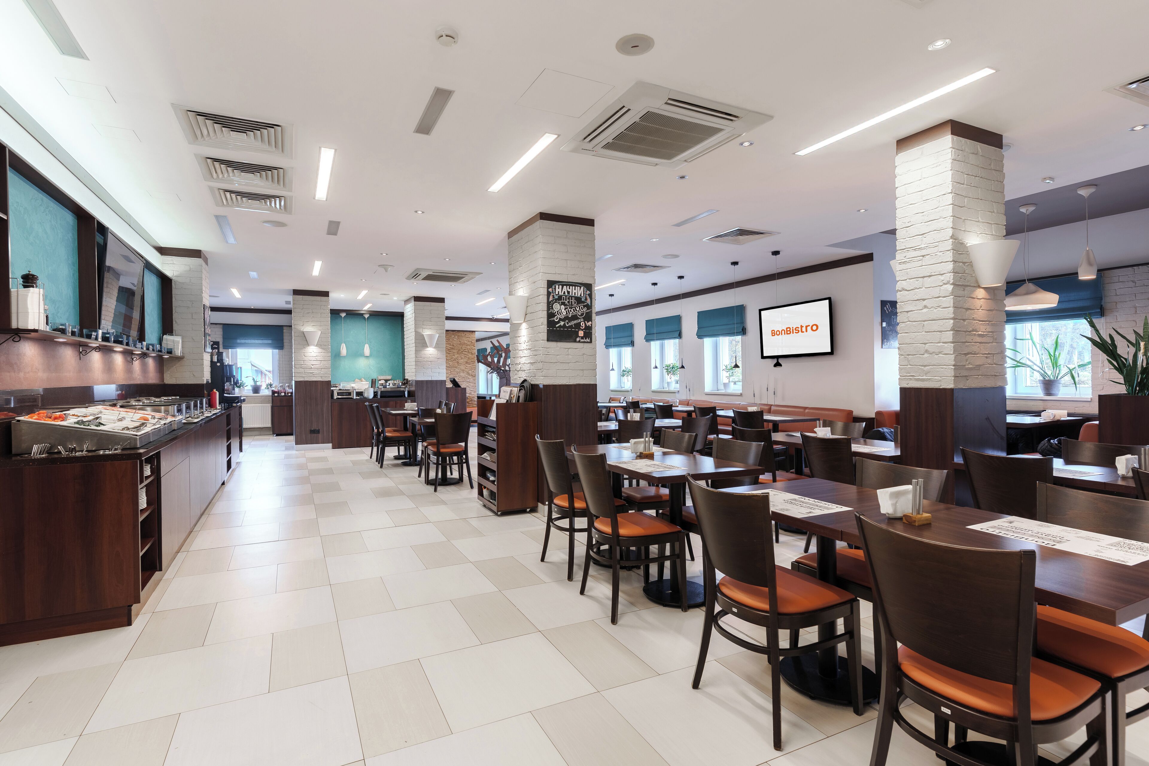 A spacious restaurant with high tables and chairs, a buffet area, and a large screen displaying the restaurant's name.