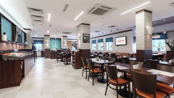 A spacious restaurant with high tables and chairs, a buffet area, and a large screen displaying the restaurant's name.