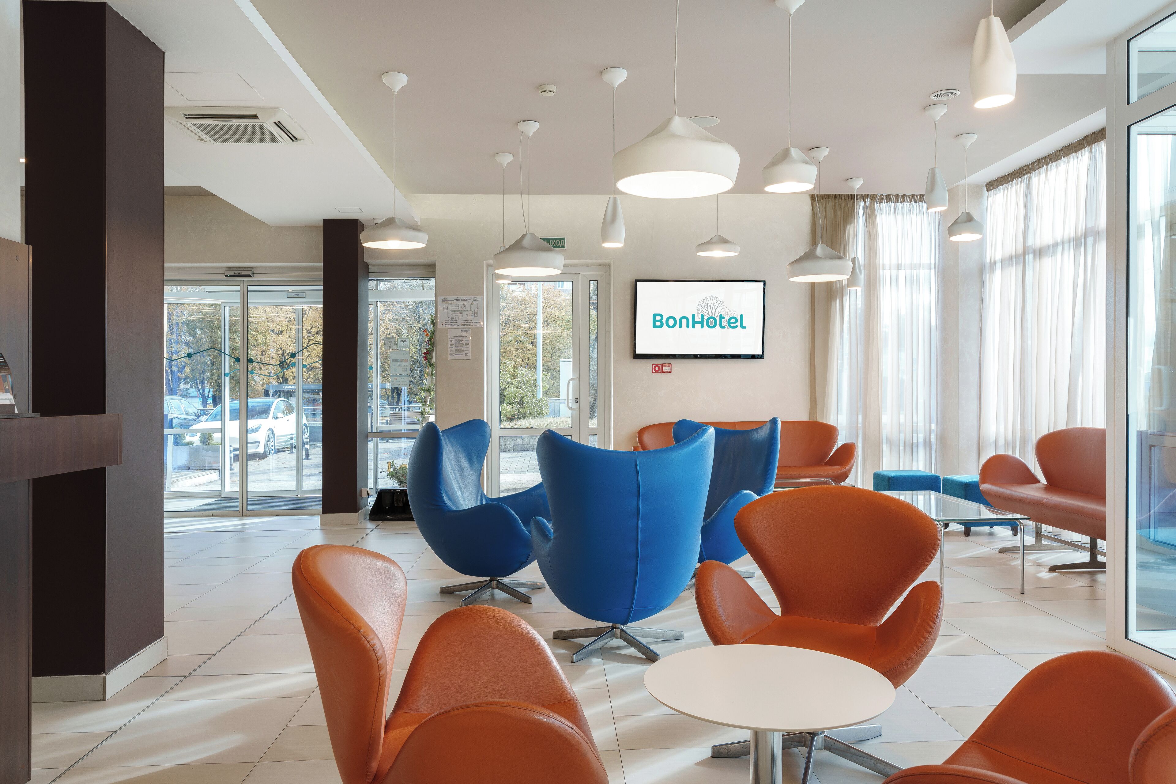 Eine Hotelhalle mit modernen, orangefarbenen Sesseln, einem weißen Tisch und einem großen Bildschirm, der den Hotelnamen zeigt.