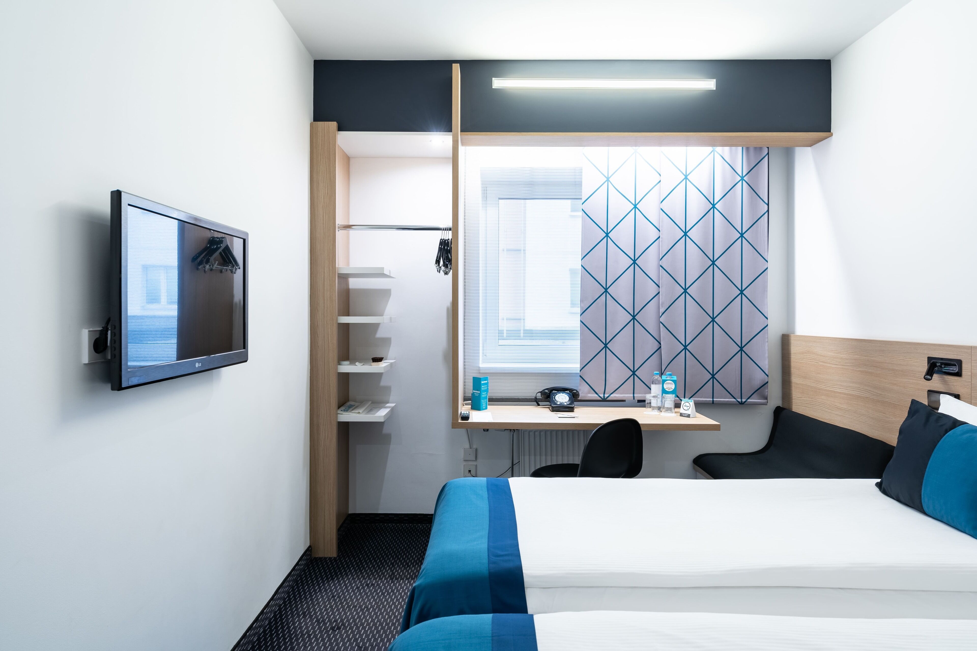 A hotel room with a bed, a desk with a computer, a TV, and a window with a geometric pattern.