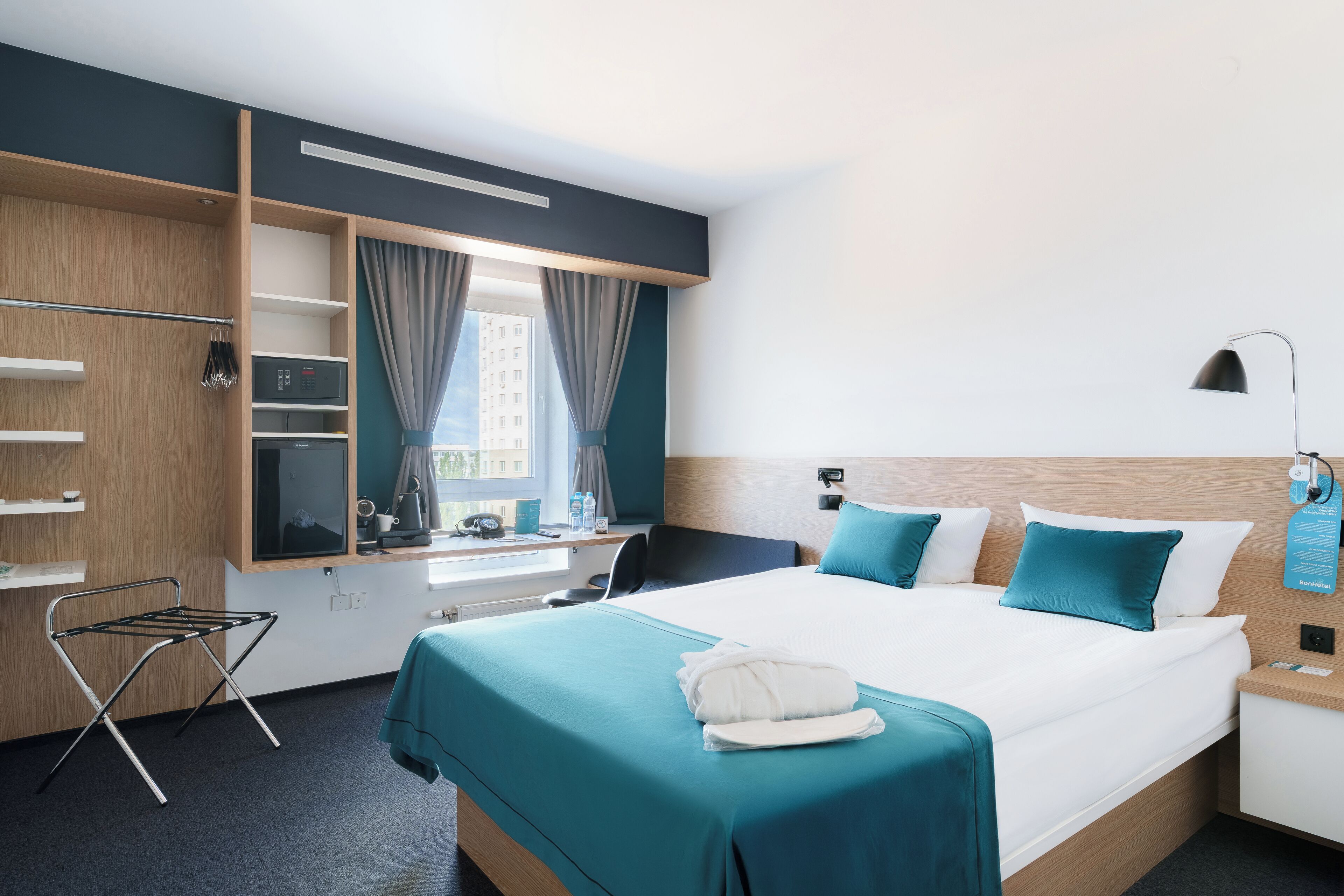 A hotel room with a bed, a desk, a chair, a small table, a lamp, a window with curtains, and a towel on the bed.