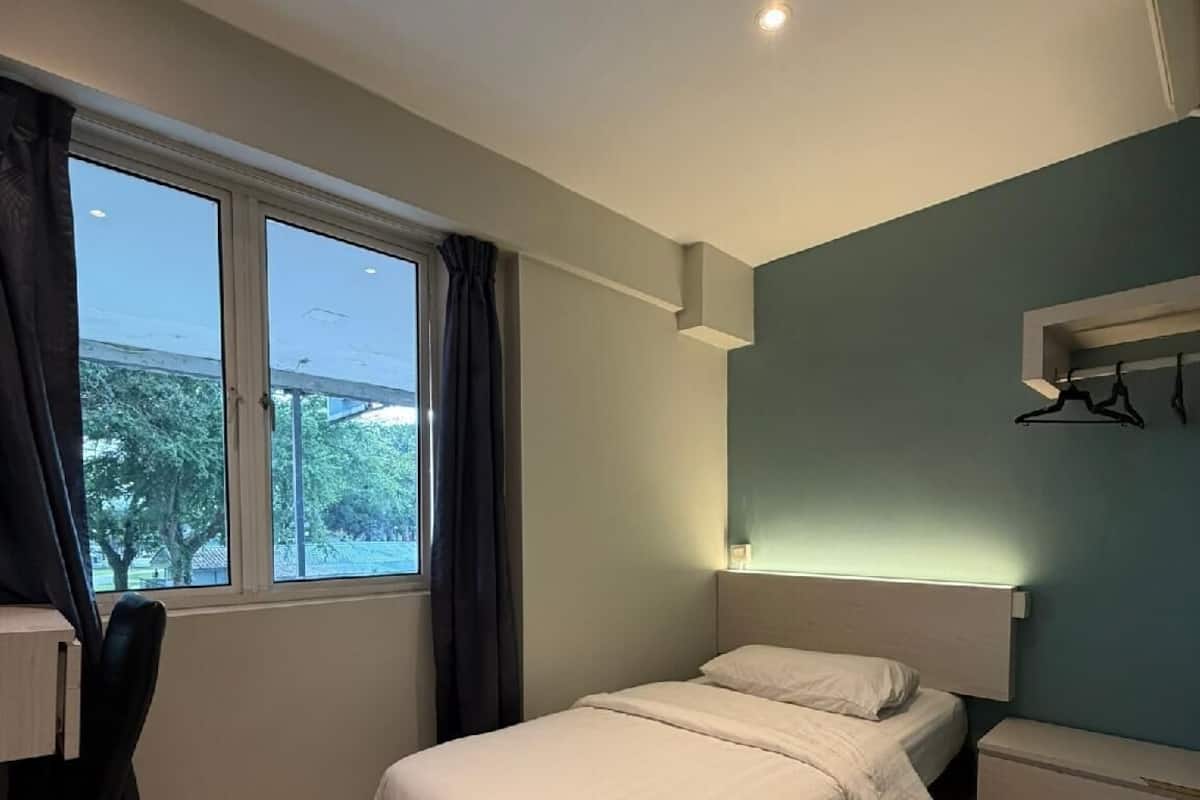 A bedroom with a bed, a desk, a chair, a window with curtains, and a wall-mounted shelf.