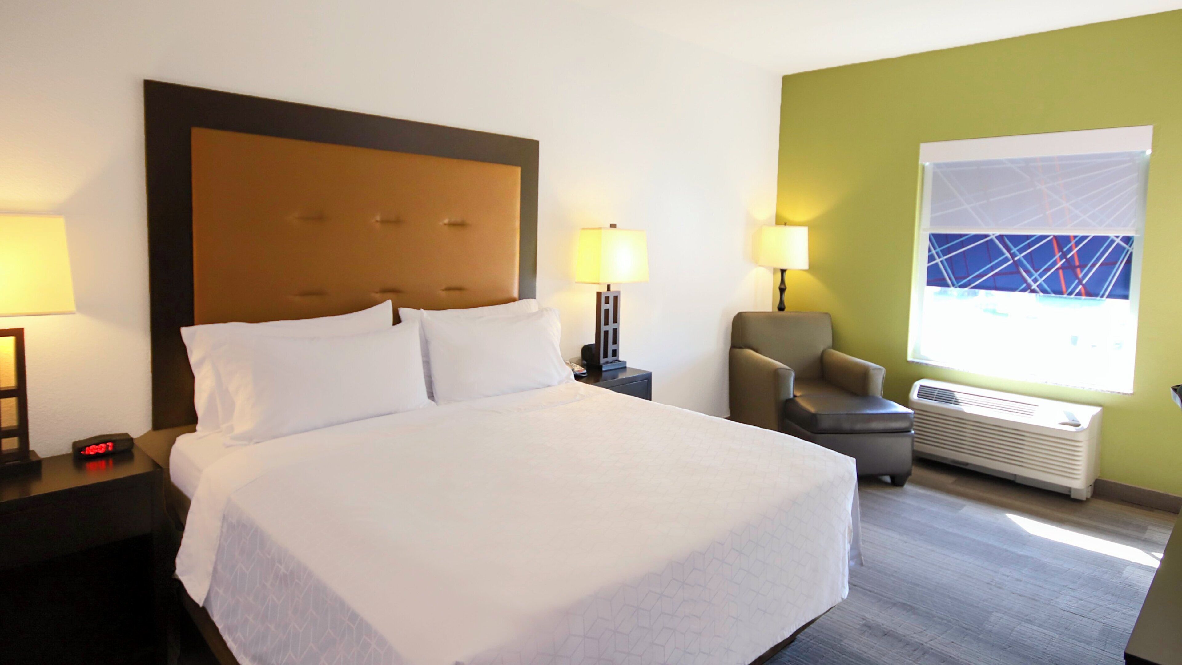 A hotel room with a large bed, two bedside lamps, a chair, a window with a view, and an air conditioning unit.