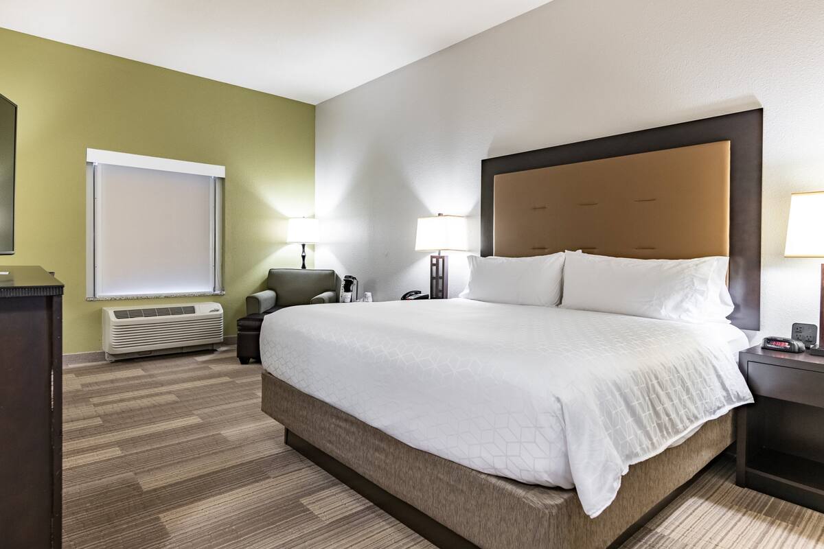A hotel room with a large bed, a desk, a chair, a lamp, and a window with blinds.