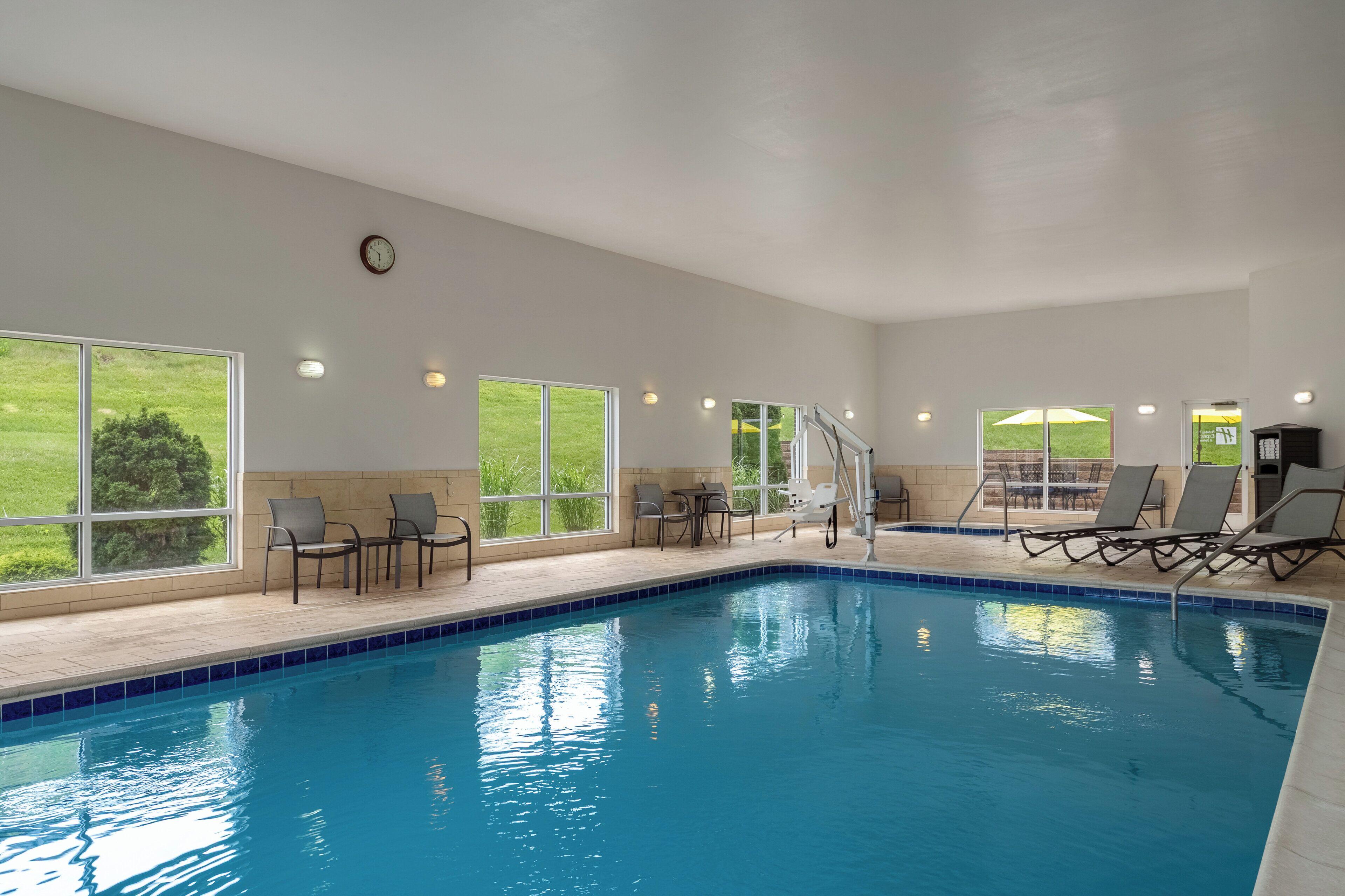 An indoor swimming pool with clear views of the outdoors, surrounded by lounge chairs and tables.