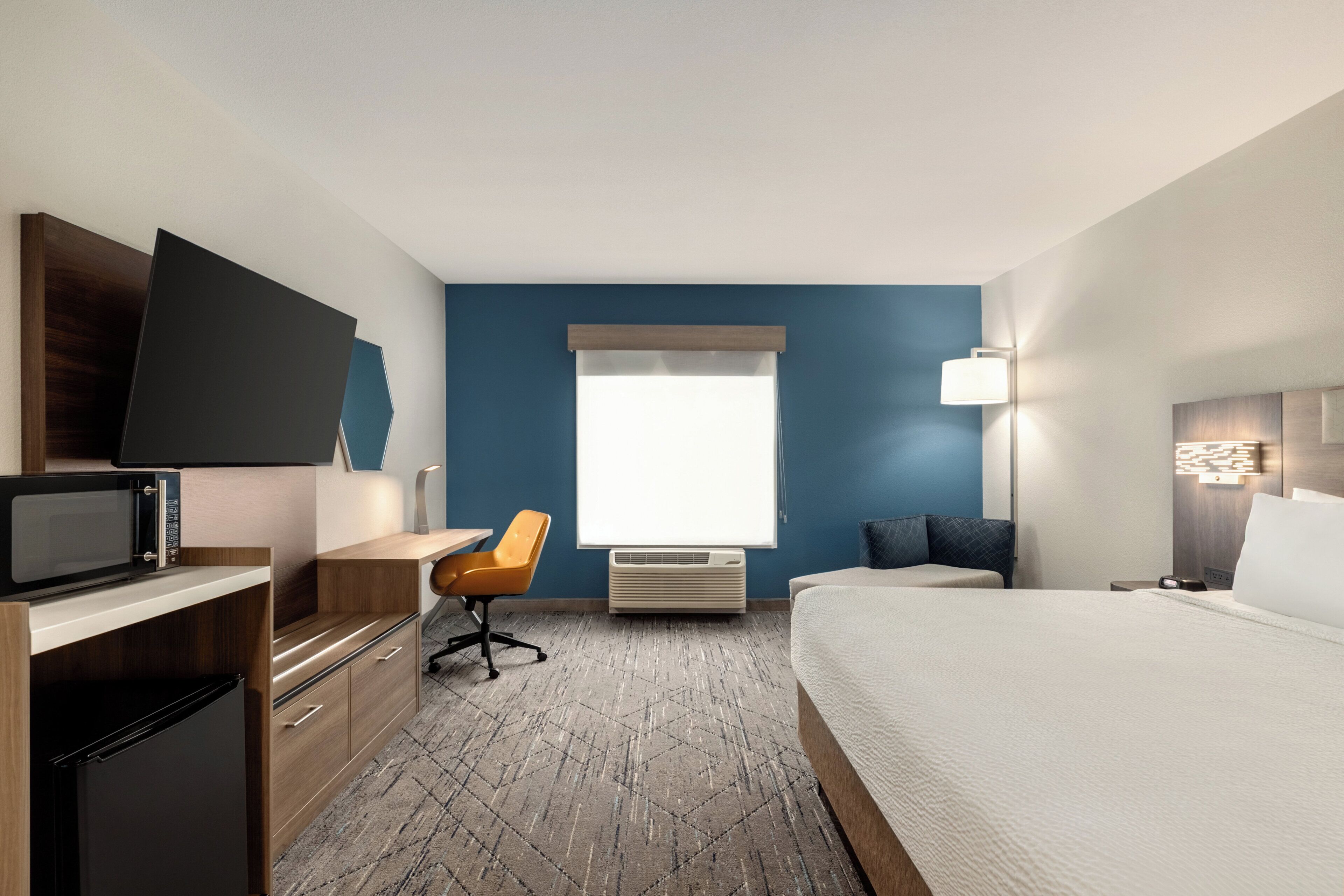 Habitación de hotel con cama, escritorio, silla, televisión de pantalla plana, microondas, aire acondicionado y ventana con persianas.