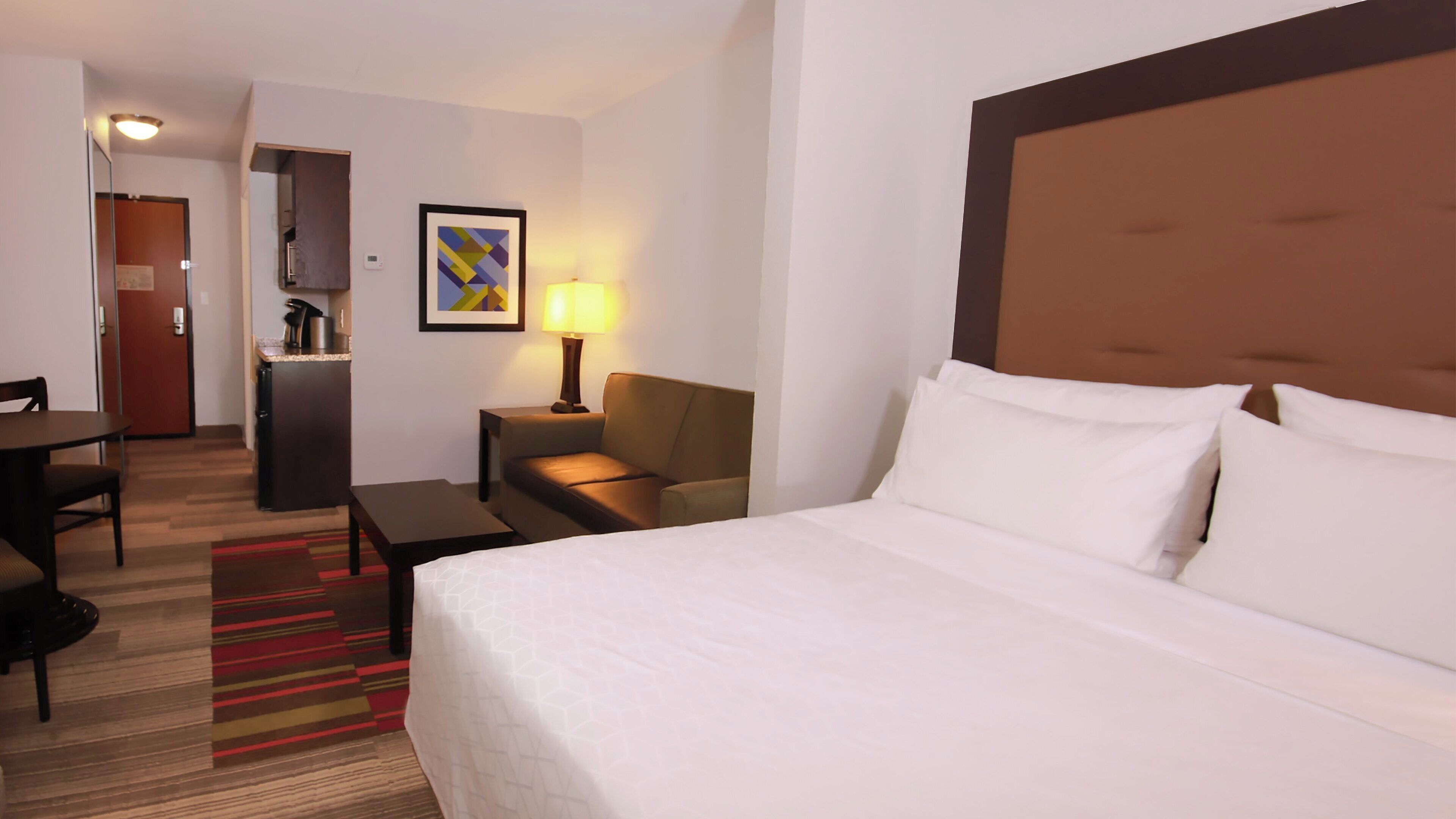 A hotel room with a bed, a desk, a chair, a lamp, and a painting on the wall.