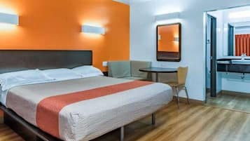 A hotel room with a bed, a small table, a chair, and a bathroom visible through an open door.