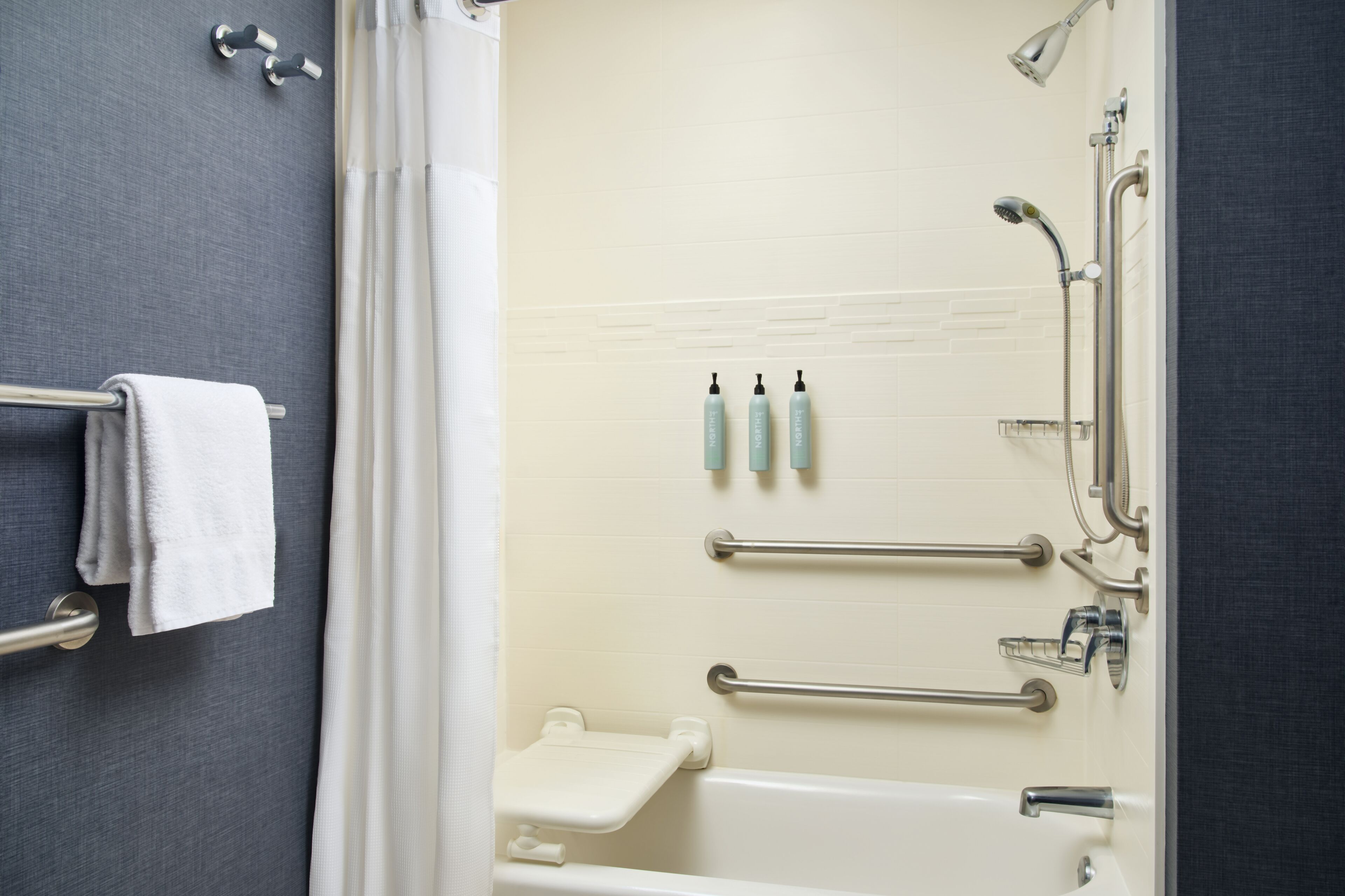 Un baño con bañera, ducha y retrete. Hay barras de apoyo y un perchero para toallas.