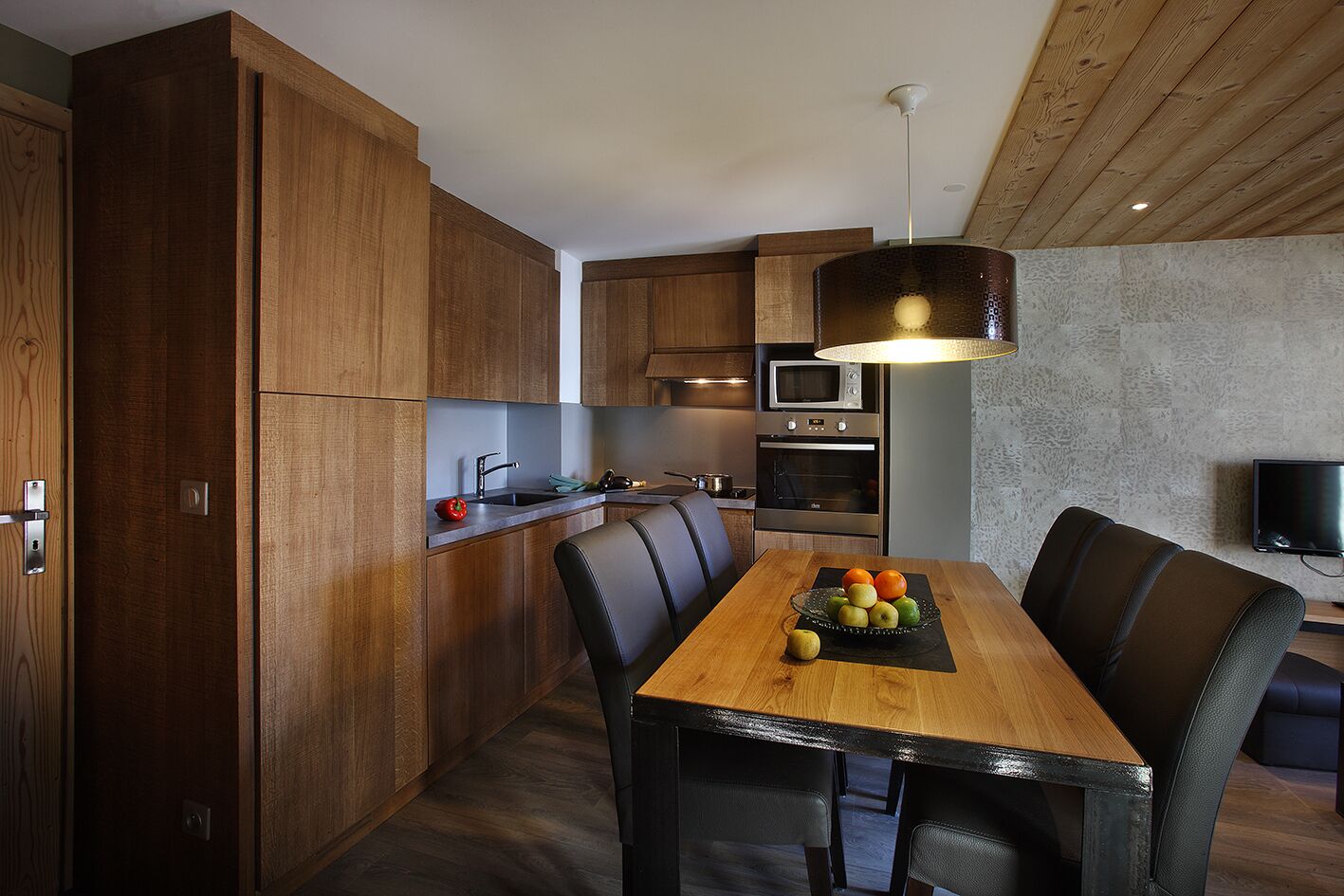A modern kitchen with wooden cabinets, a dining table with chairs, a fruit bowl, and a television.