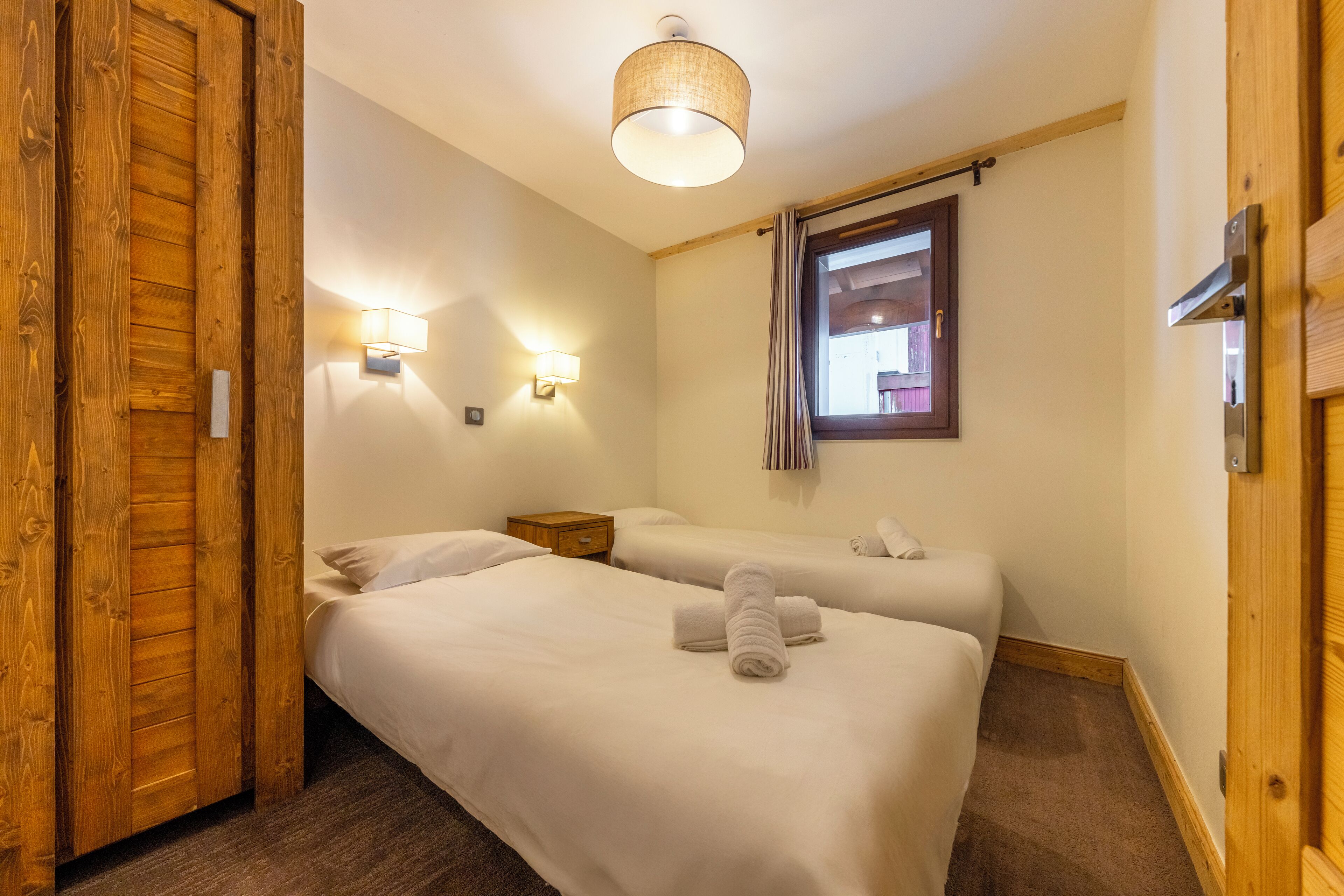 Een hotelkamer met twee bedden, een houten deur, een klein nachtkastje van hout, een wandlamp en een raam met gordijnen.