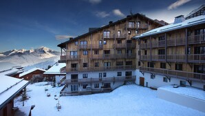 Un bâtiment en bois à plusieurs étages, doté de balcons, une cour enneigée et une vue sur les montagnes en arrière-plan.