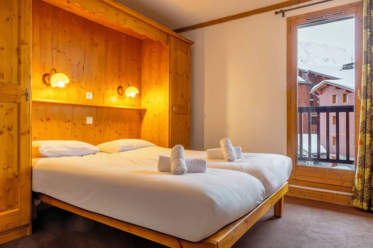 Une pièce en bois avec deux lits, chacun étant équipé de draps et de serviettes blancs. Des lampes murales sont fixées au-dessus des lits, et une fenêtre offre une vue sur les bâtiments à l’extérieur.