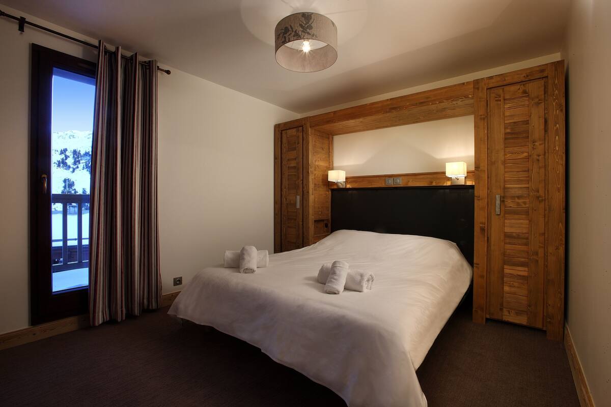 Une chambre d’hôtel avec un grand lit, une tête de lit en bois, deux lampes de chevet et une vue sur un paysage enneigé par la fenêtre.