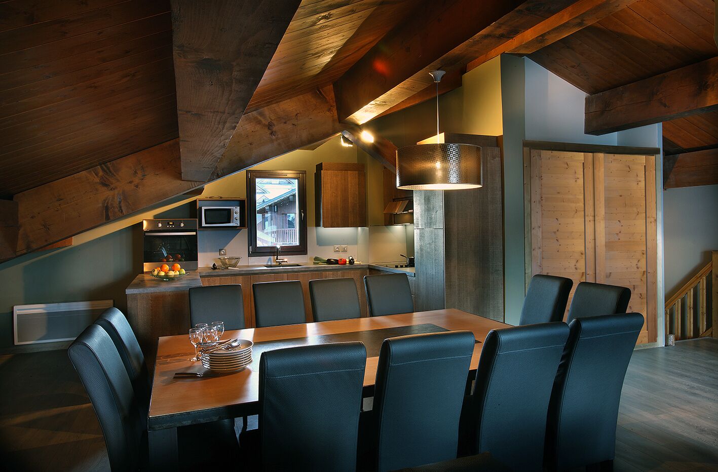 A modern kitchen with a dining area, featuring wooden beams, a large window, and a view of the outdoors.
