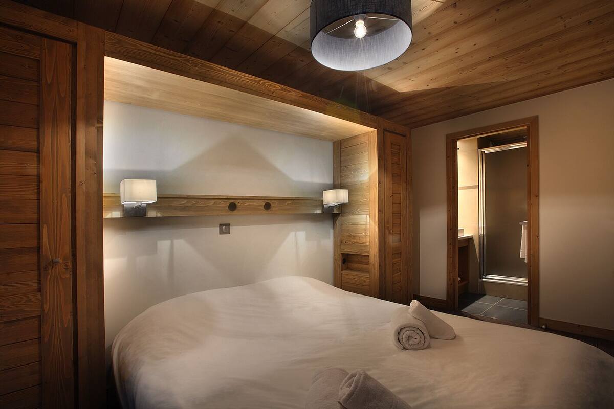 Une chambre à coucher aux murs lambrés, avec un grand lit, deux lampes fixées au mur et un luminaire au plafond.
