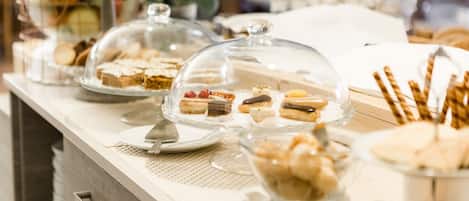 Una mesa de buffet con diversos postres bajo campanas de cristal, como pasteles, bollería y fruta.