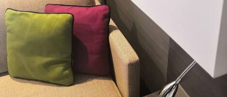 A modern hotel room with a beige sofa, two pillows, a wooden side table, and a white lamp.