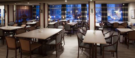 A spacious dining area with multiple tables and chairs, large windows with curtains, and a view of the street outside.