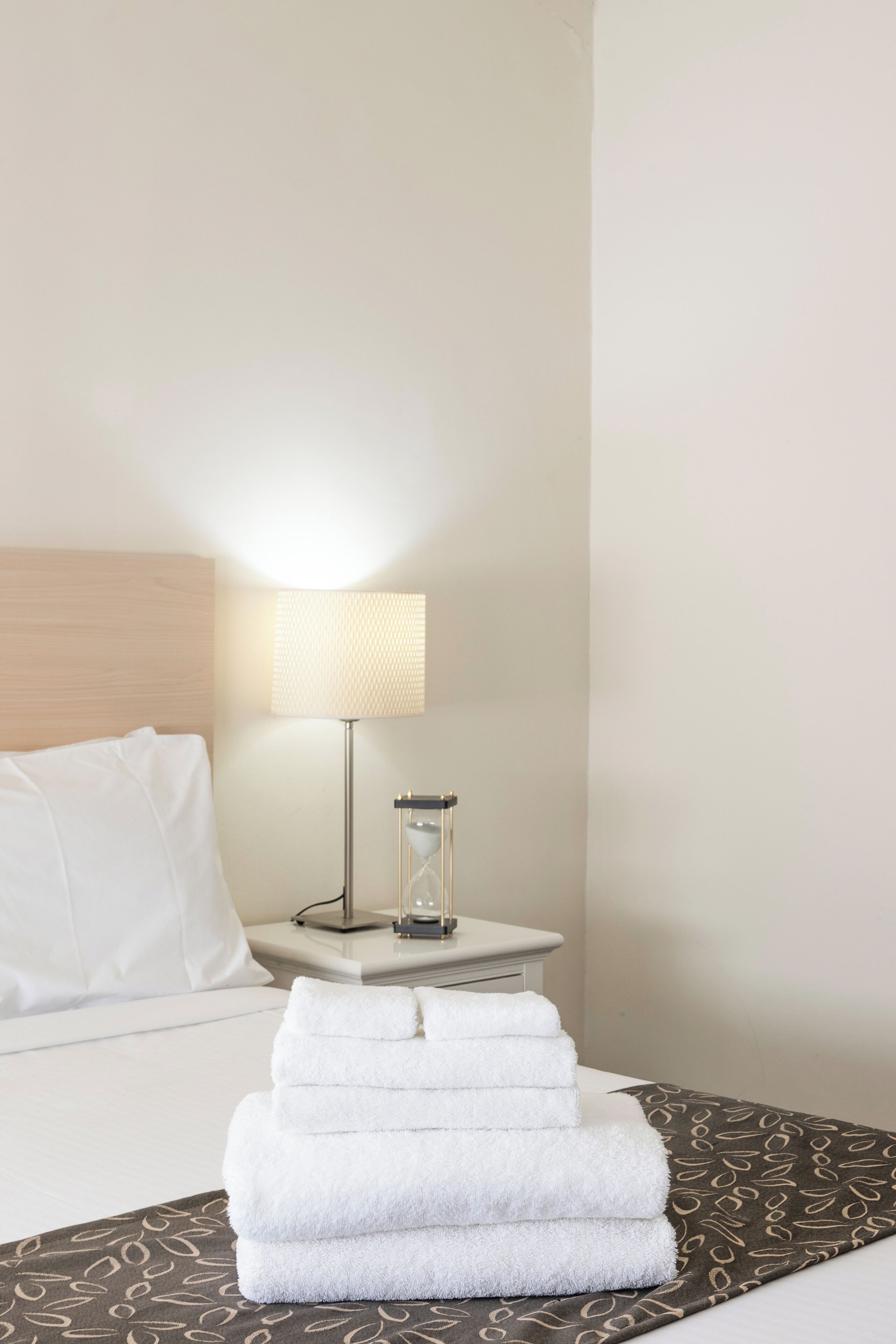 Una cama bien hecha con sábanas blancas y una colcha marrón estampada. Un montón de toallas blancas está sobre la cama. Se ve una mesita de noche con una lámpara y un recipiente de vidrio.