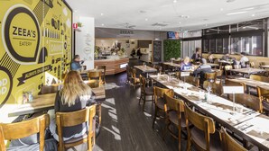 Interior moderno de un restaurante con mesas y sillas de madera, una zona de bar y una pared amarilla con el rótulo 'ZEE'S EATERY'.