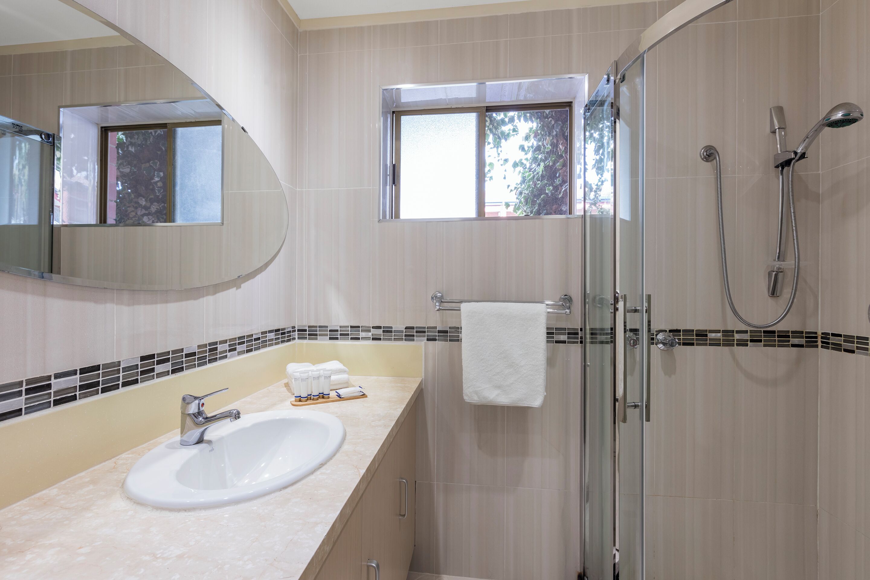 A bathroom with a sink, mirror, shower, and a window.