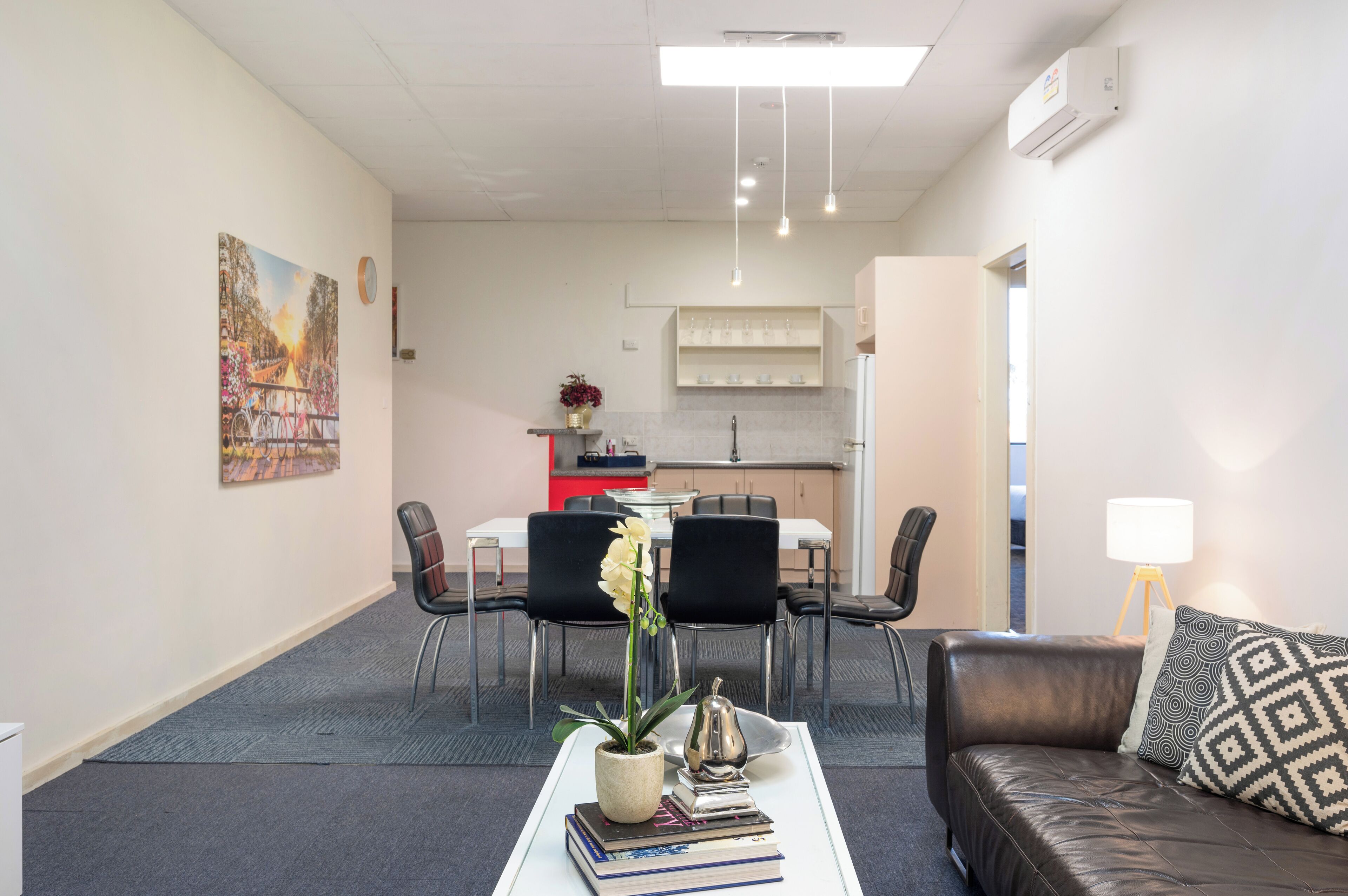 Una sala moderna con un sofá de cuero, una mesa de centro con libros y un jarrón, un área de comedor con sillas y una mesa, y una cocina al fondo.