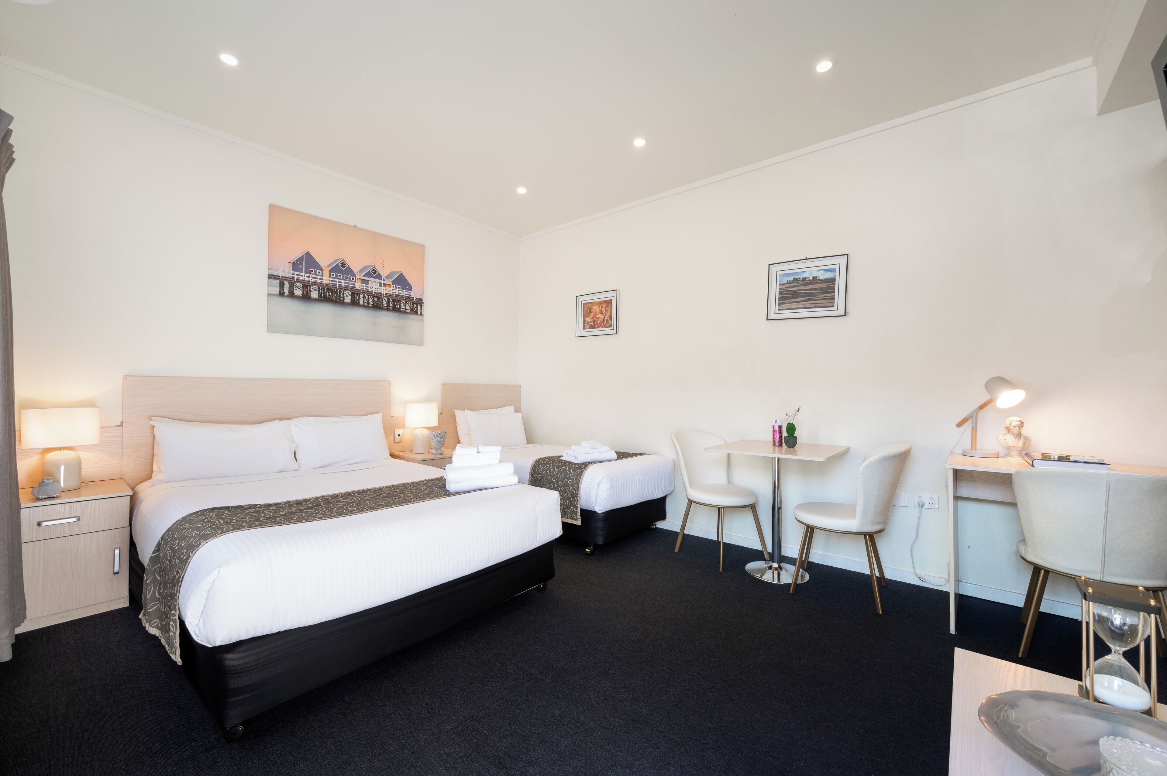 A hotel room with two beds, a desk, two chairs, a small table, and a painting on the wall.