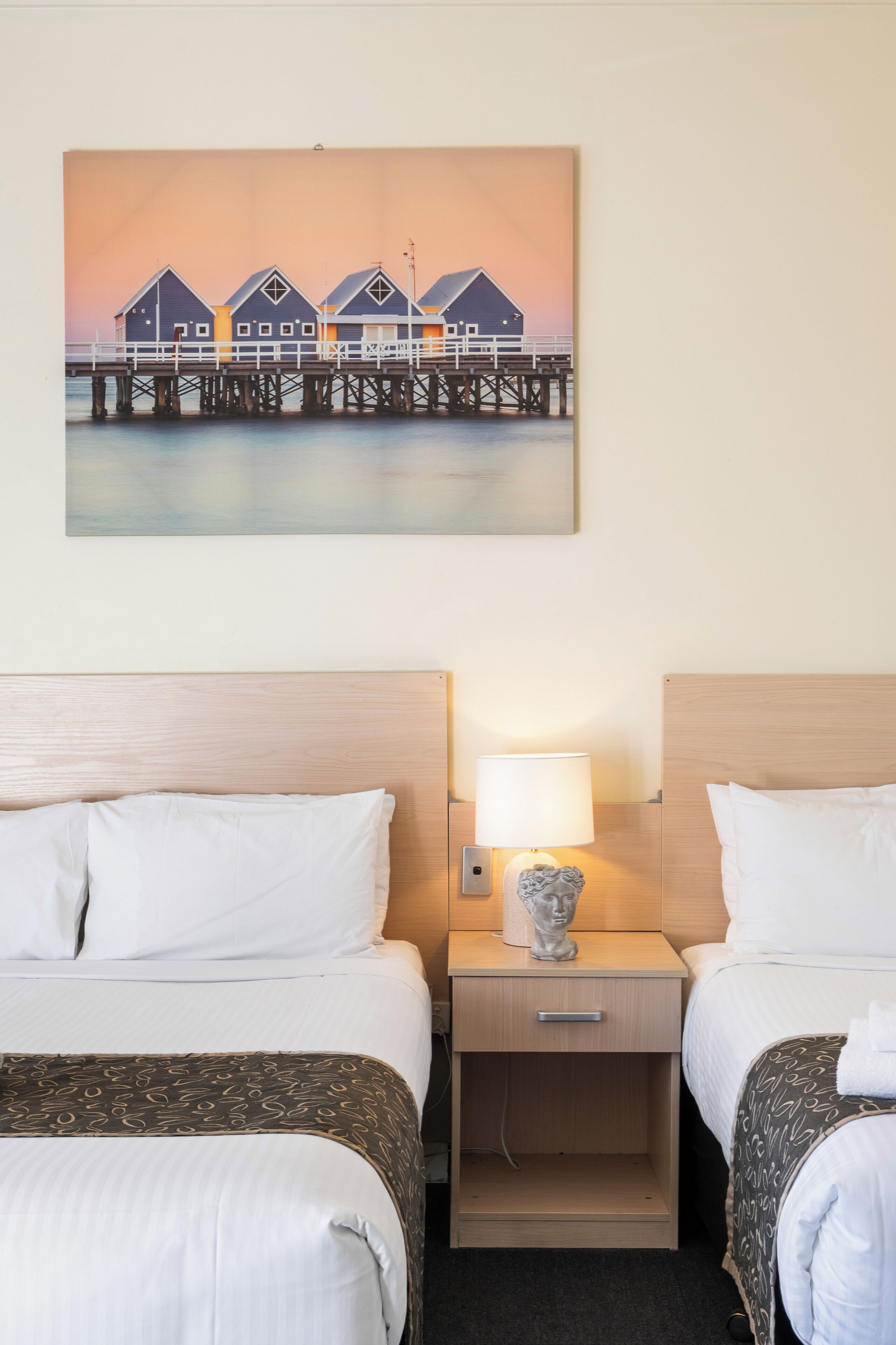 A hotel room with two beds, a nightstand with a lamp, and a painting of a pier.